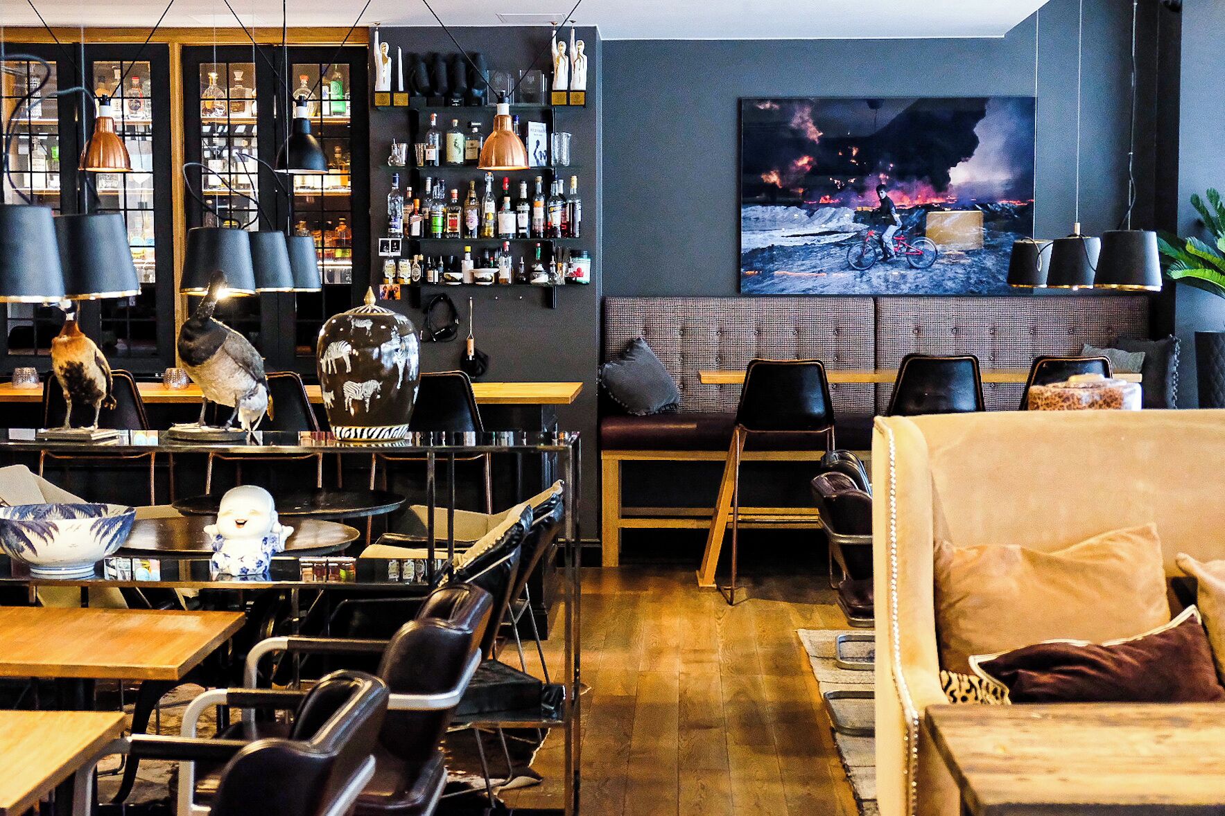 Uma área de bar com prateleiras de garrafas, uma pintura grande de um ciclista e um espaço de estar com sofá e poltronas.