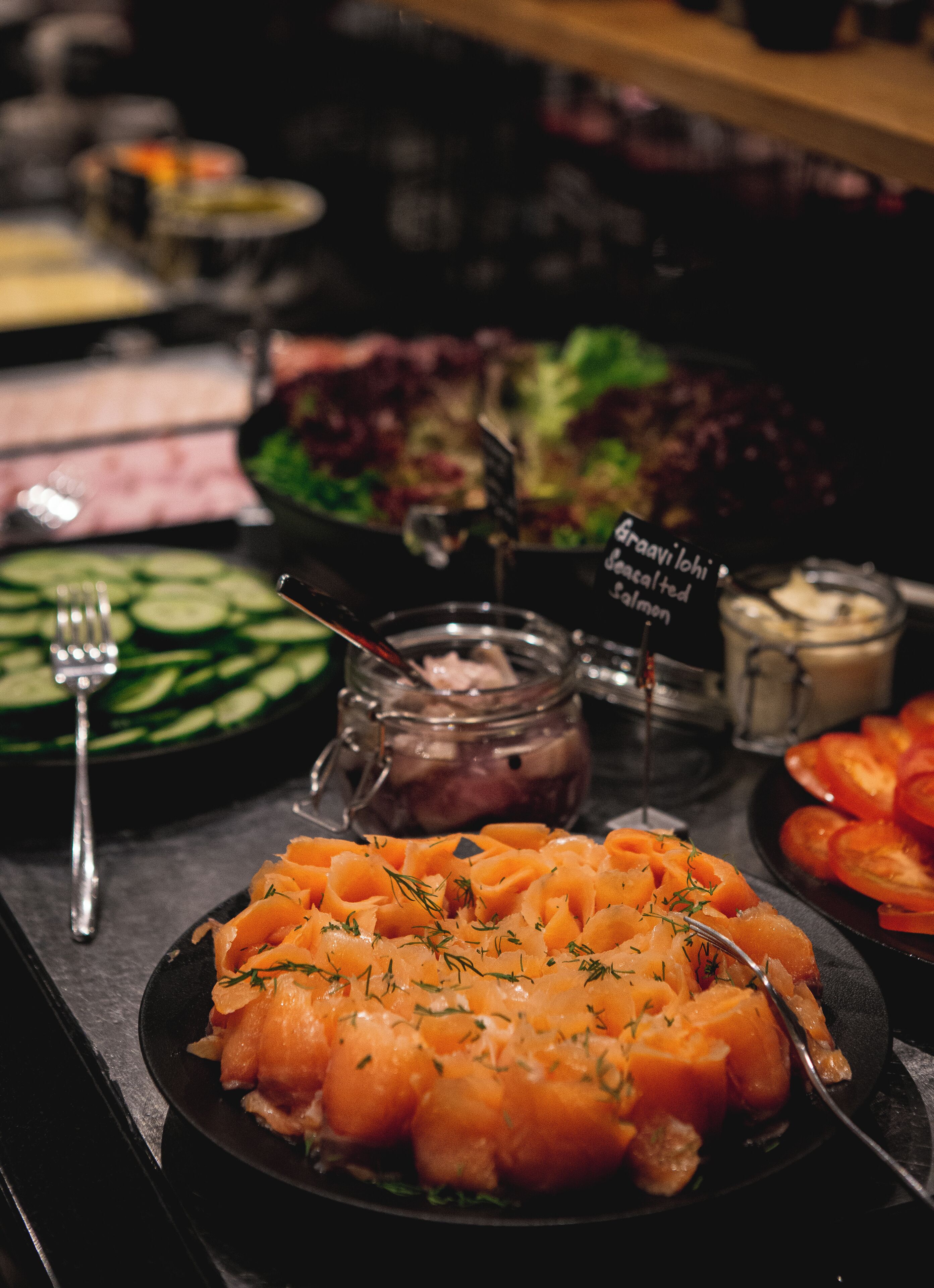 A buffet with various dishes including salmon, salad, and sliced vegetables.