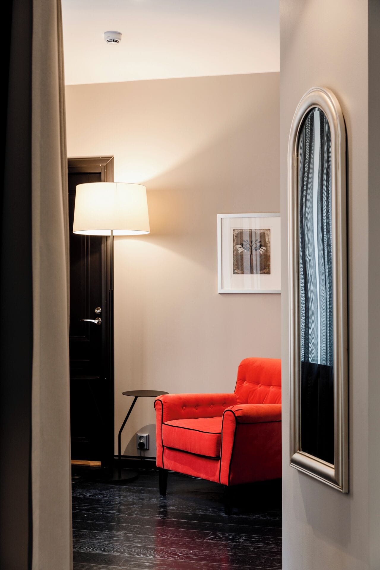 Een hal met een rode fauteuil, een vloerlamp, een ingelijst schilderij aan de muur en een deur met een lichtschakelaar.