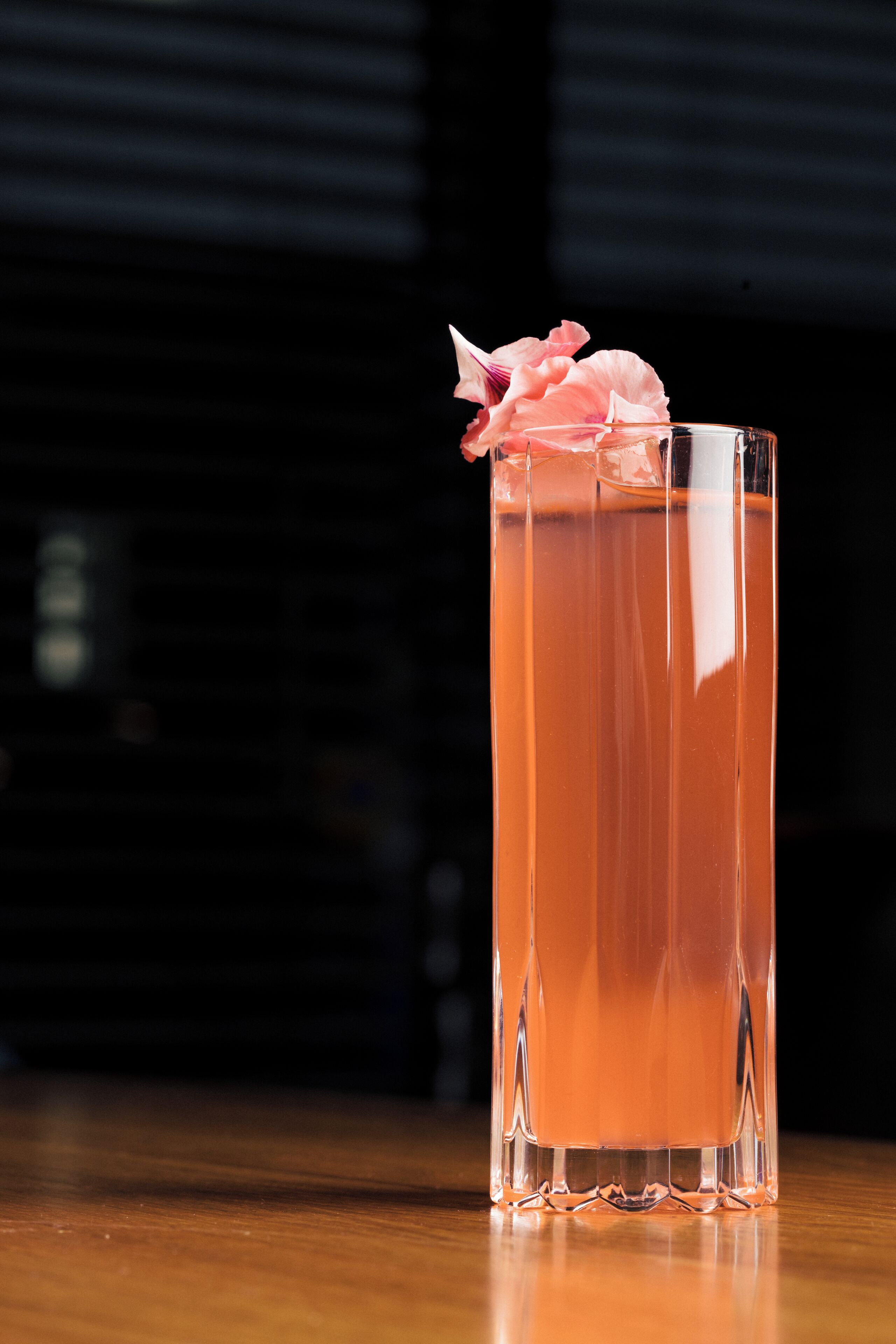 A tall glass with a pink drink and a flower garnish on a wooden surface.