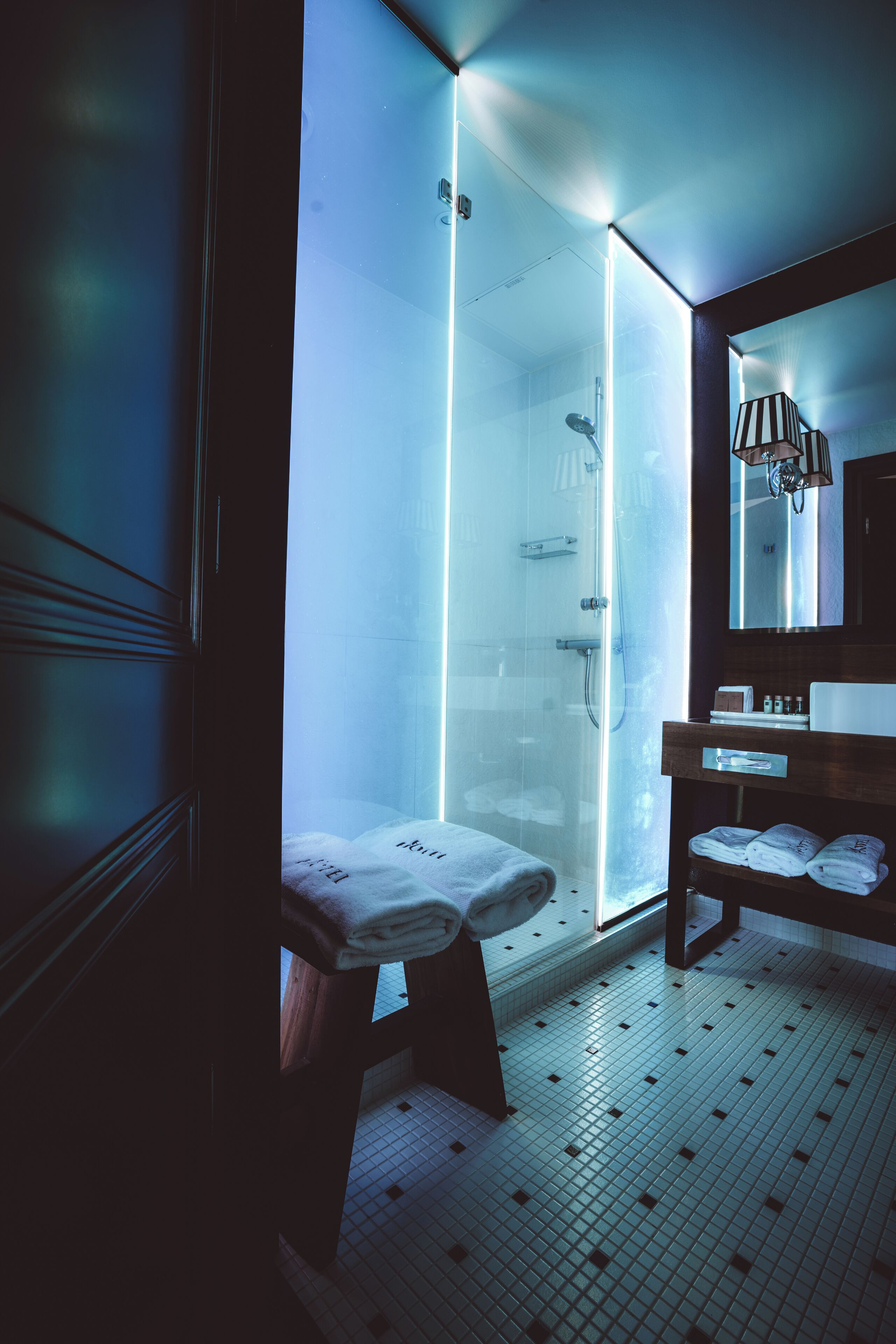 A modern bathroom with a glass shower enclosure, a wooden bench, a sink, and a mirror with a striped lampshade.