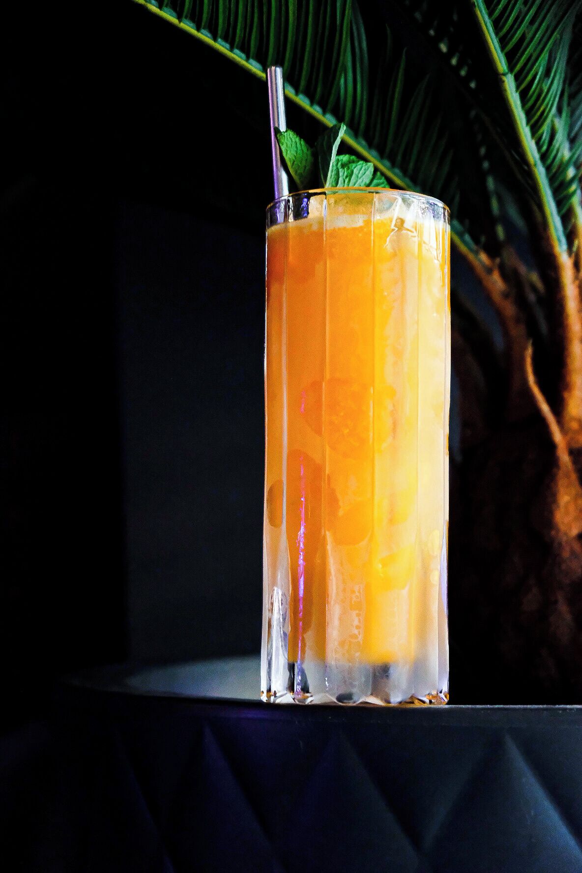 A tall glass with a straw and mint garnish, placed on a dark surface with a palm leaf in the background.