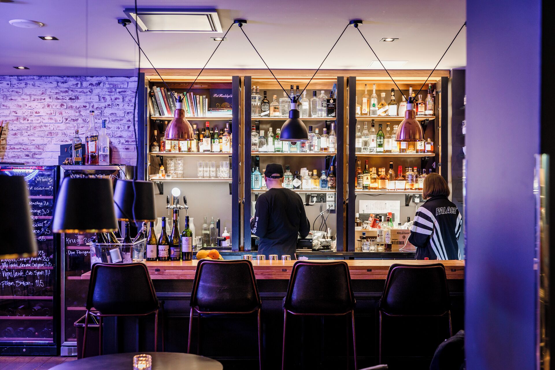 Un bar con pared de ladrillo, estantes llenos de botellas de licor y taburetes.
