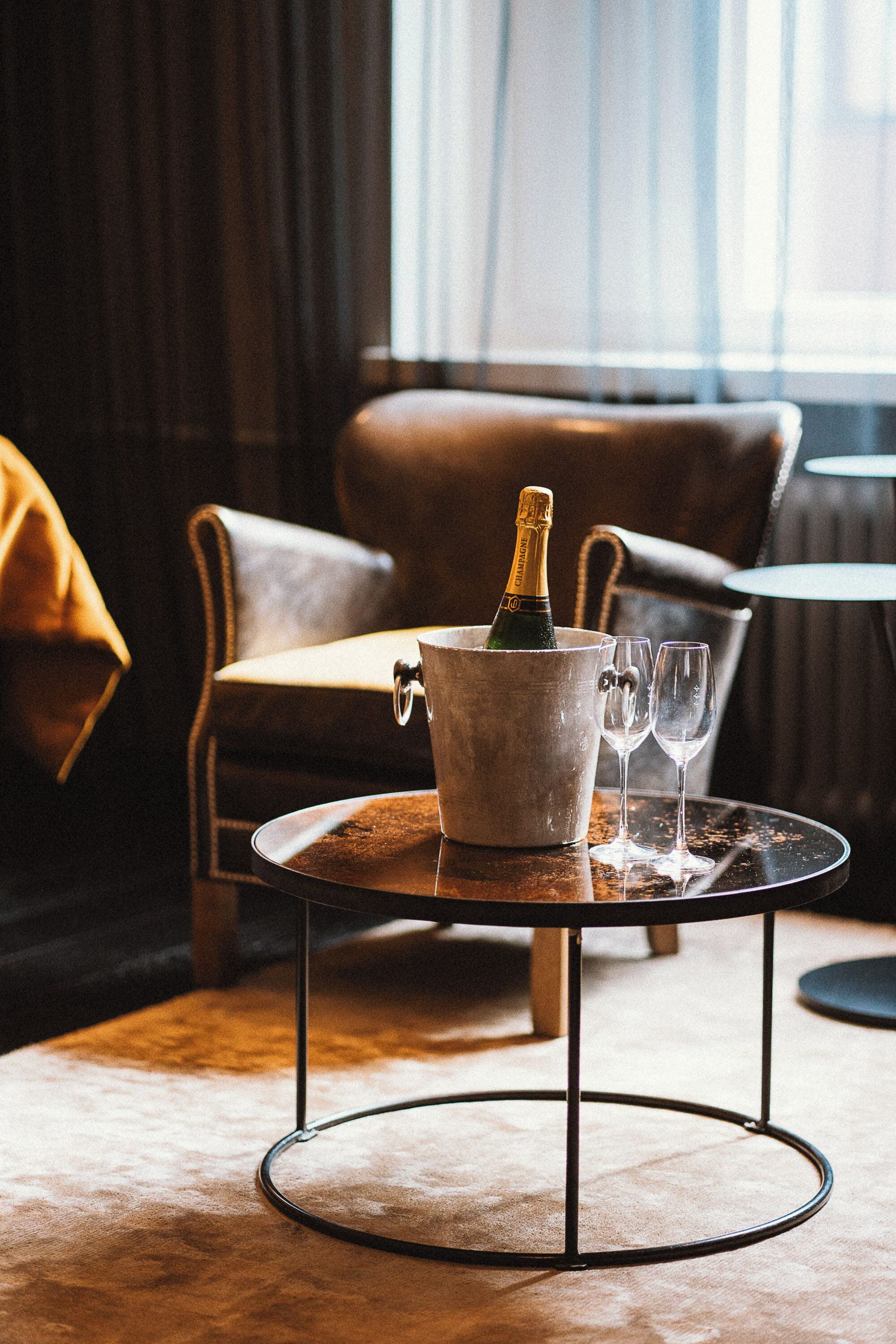 Una habitación con un sillón de cuero, una mesita redonda con base metálica, una botella de champán y dos copas de vino.