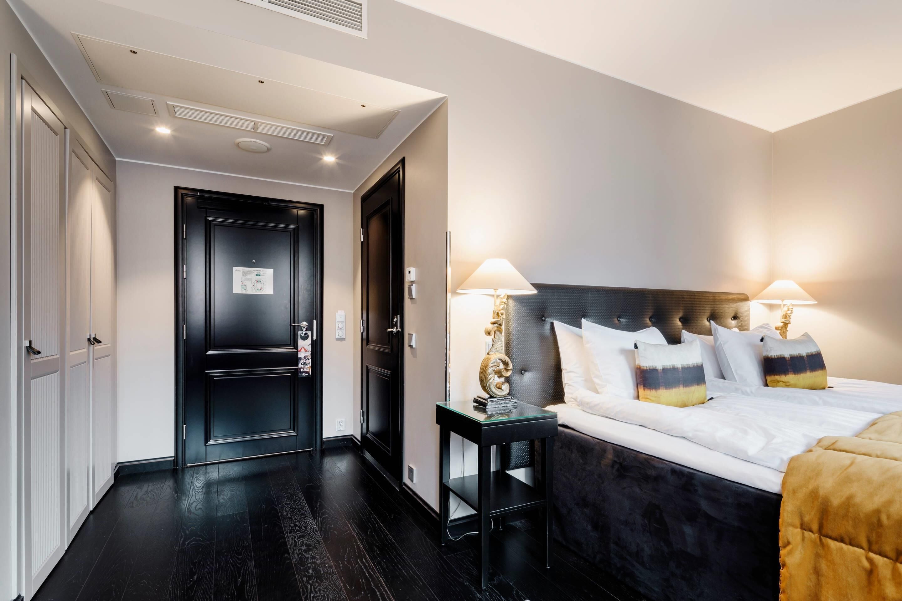 A hotel room with a dark wood floor, a bed with white and multicolored pillows, two bedside lamps, and a black headboard.
