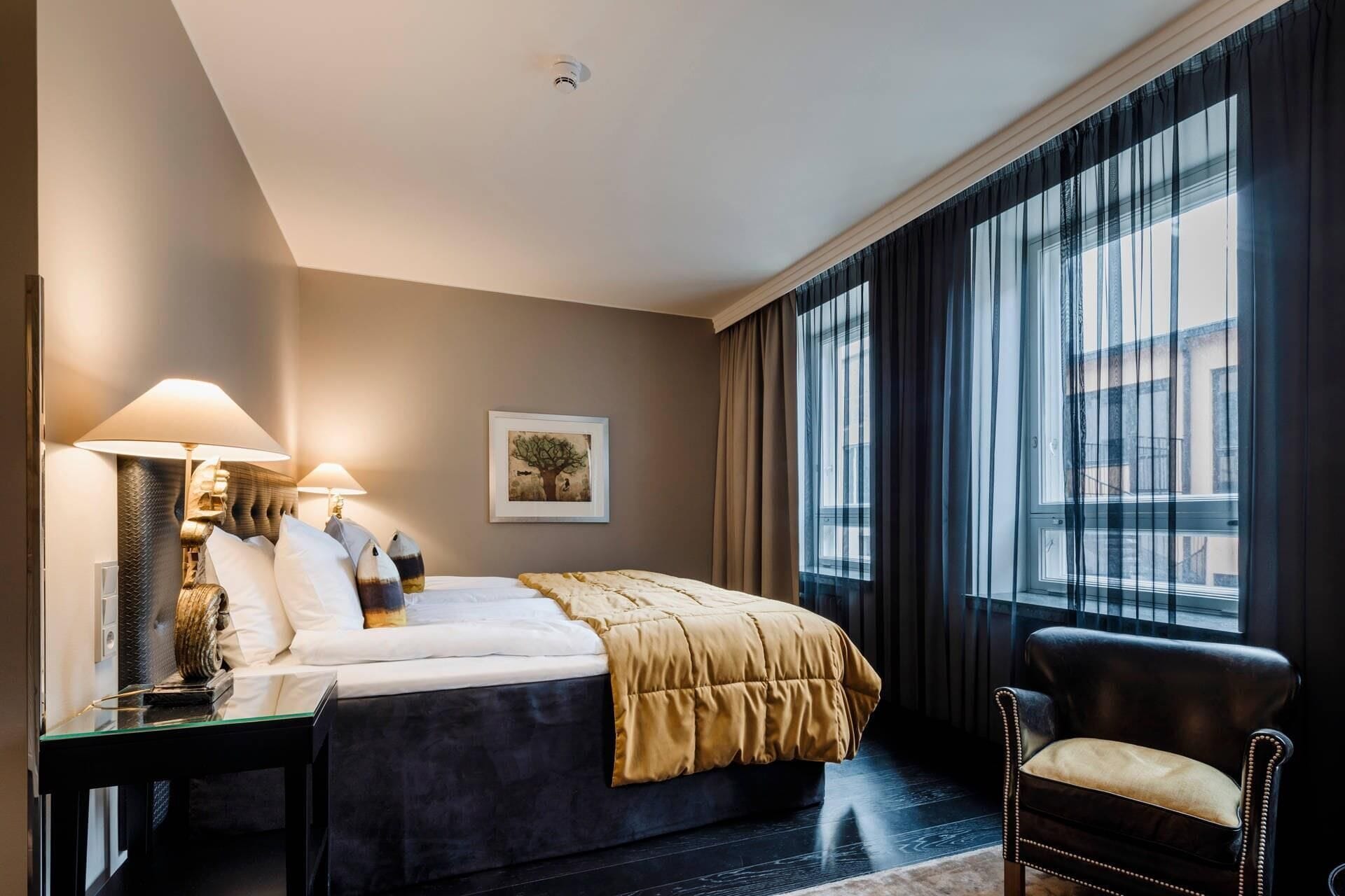 Hotellihuone, jossa on sänky, yöpöytävalaisimet, tuoli, ikkuna, jossa on verhot, ja seinällä oleva kehystetty taulu.