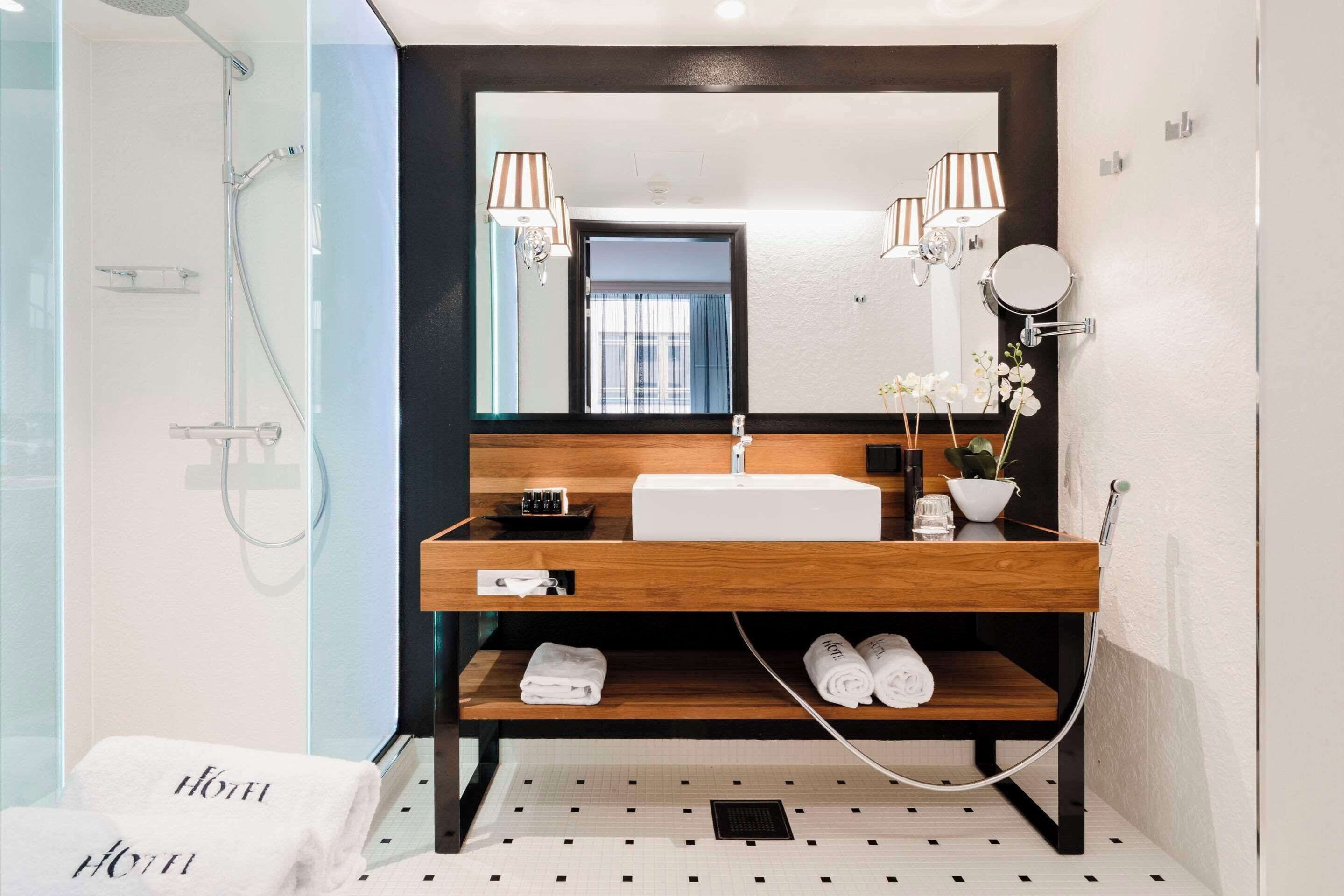 A modern bathroom with a wooden vanity, a large mirror, two wall-mounted lamps, a round mirror, a sink, and a shower area.