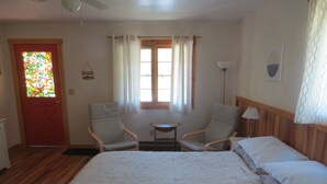 Quarto com uma cama grande, duas poltronas, uma mesinha, janela com cortinas, porta com detalhe em vitral e uma luminária.