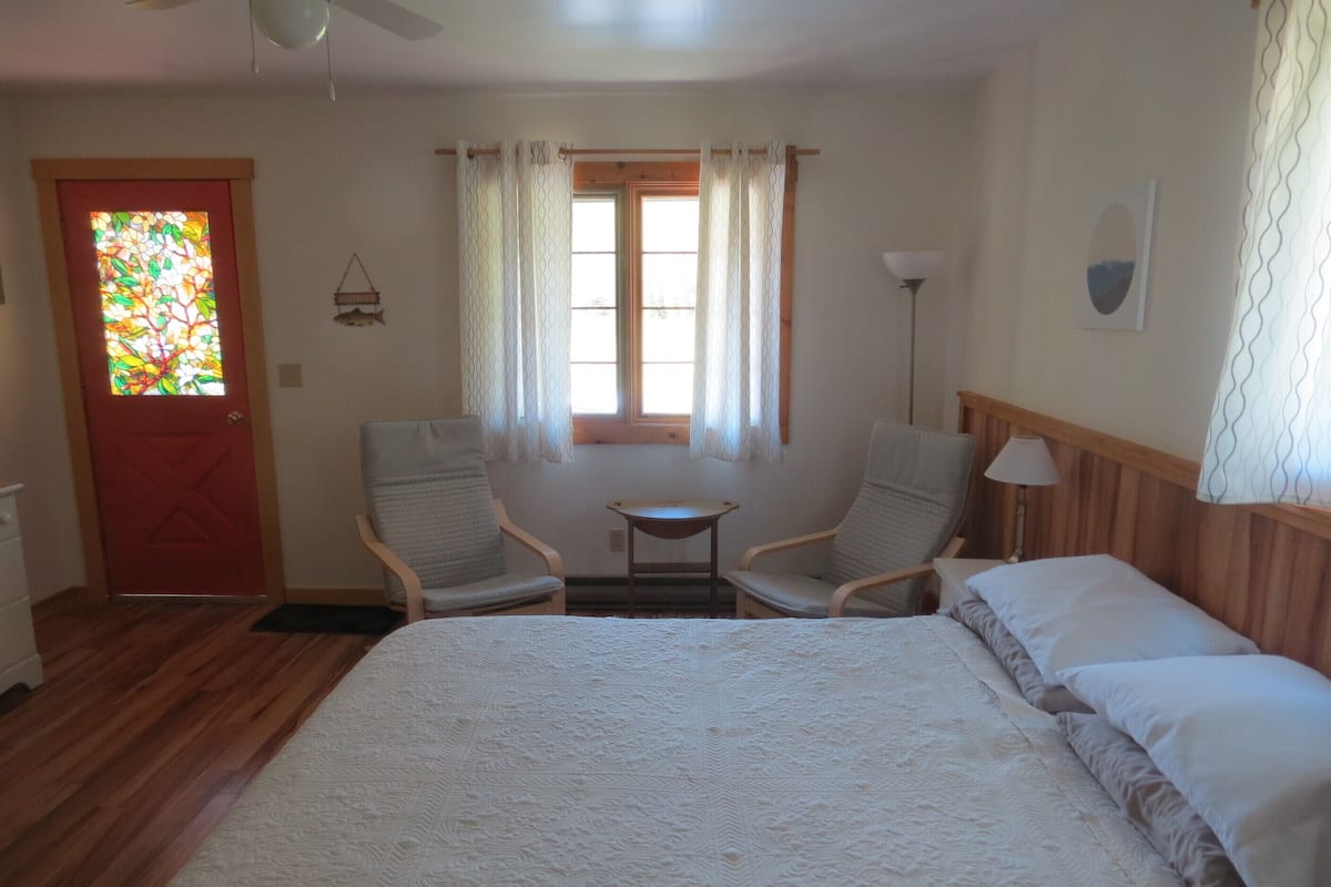 A bedroom with a large bed, two armchairs, a small table, a window with curtains, a door with a stained glass design, and a lamp.