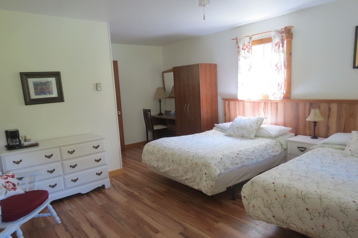 A bedroom with two beds, a wooden wardrobe, a desk with a chair, and a small table with a lamp.