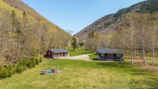Uma cabana com varanda e mesa de piquenique em área gramada, com árvores e montanhas ao fundo.