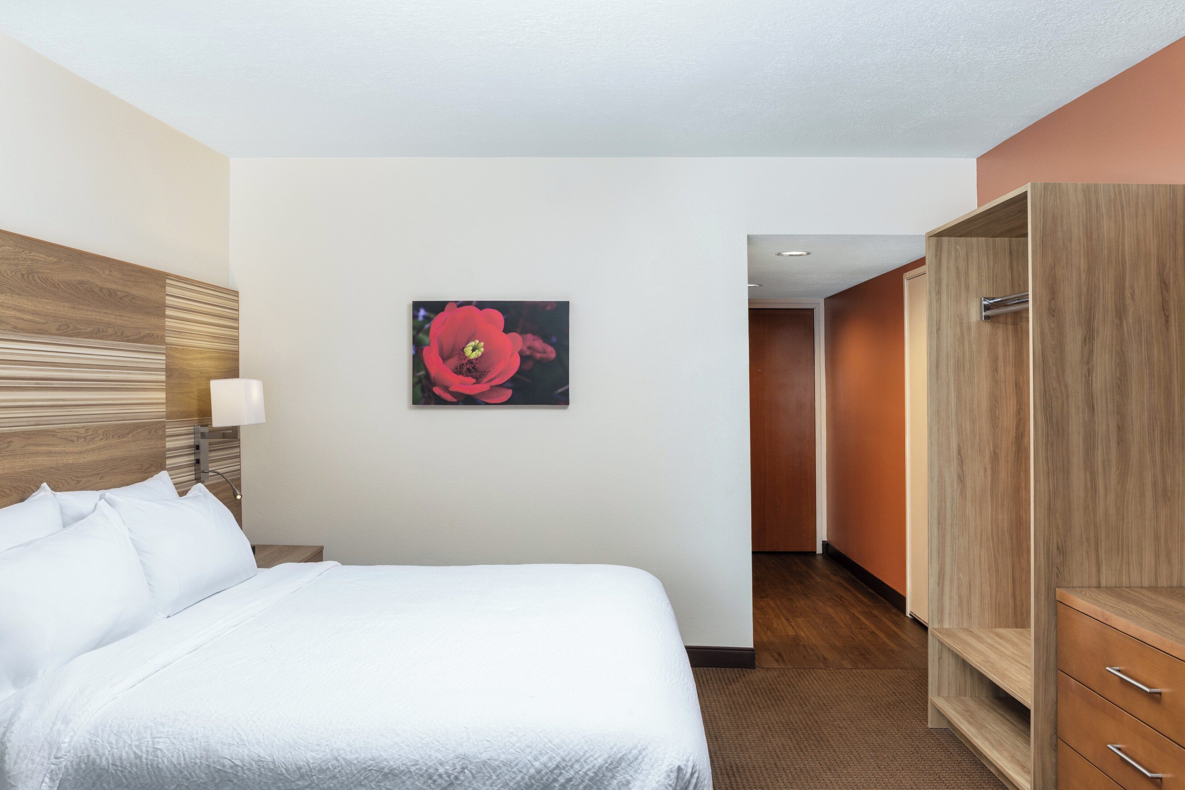 A hotel room with a bed, a wooden headboard, a nightstand with a lamp, a red flower painting on the wall, and a wooden wardrobe.