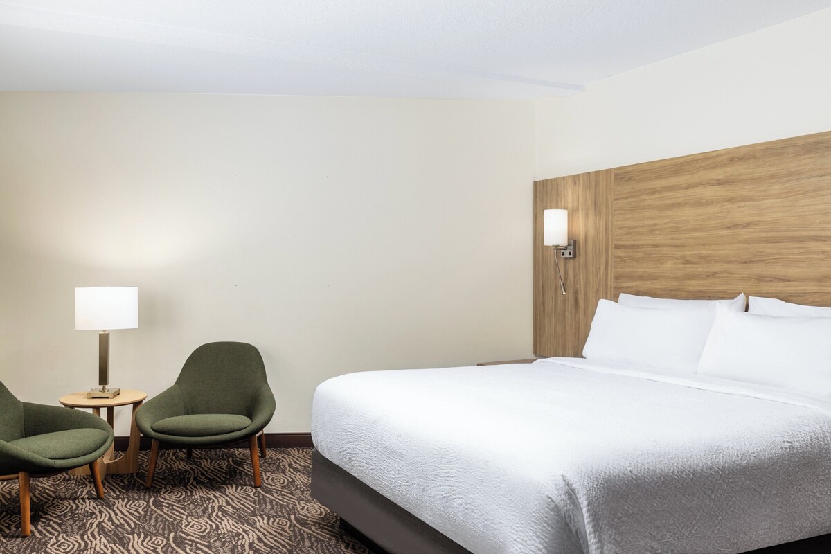 A hotel room with a large bed, two armchairs, a small table with a lamp, and a wooden headboard.