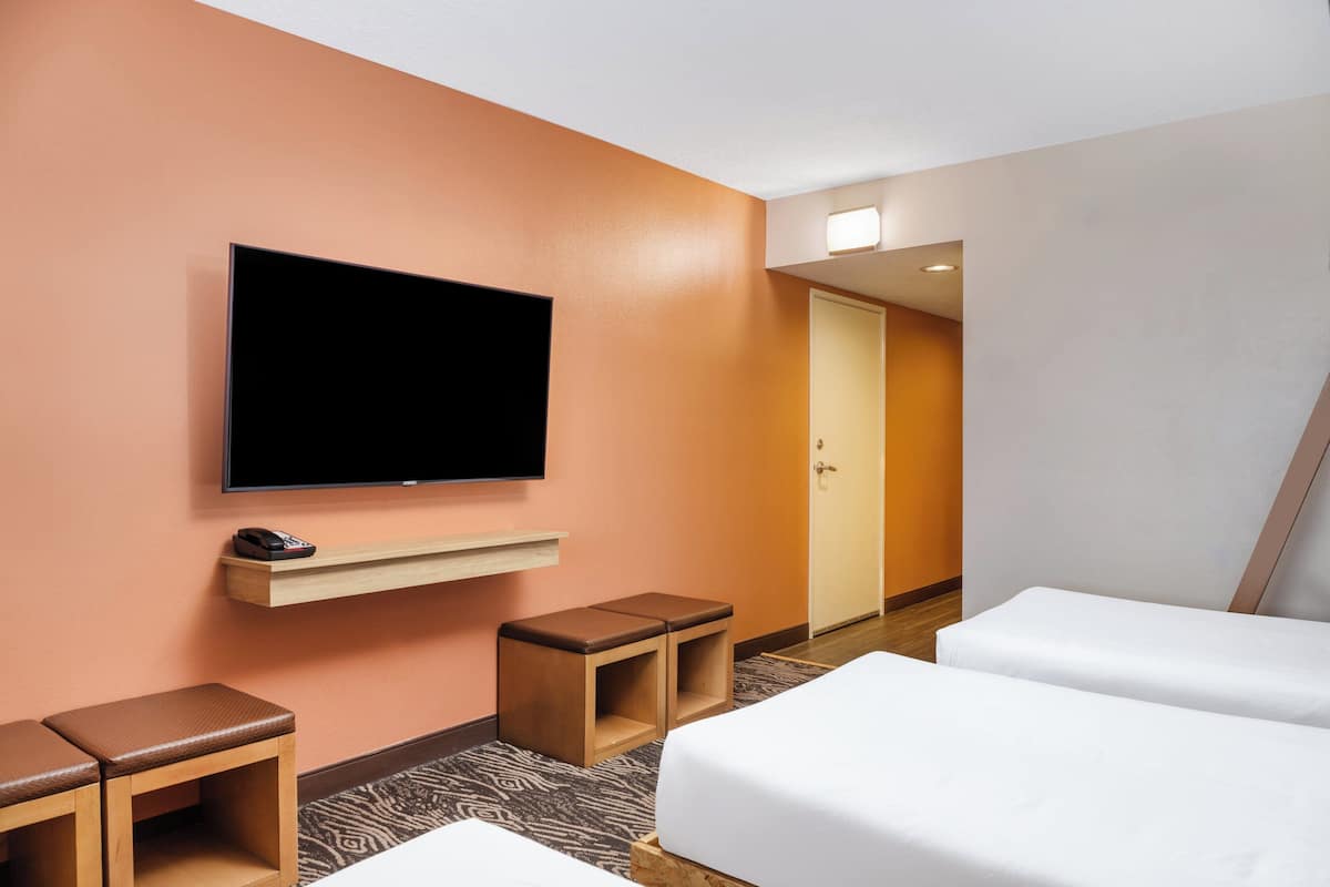 A hotel room with a flat-screen TV mounted on the wall, a wooden bench, a small wooden stool, and two beds with white bedding.