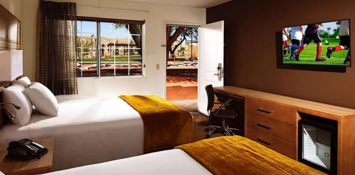 A hotel room with two beds, a desk, a chair, a TV, and a window with a view of a park.