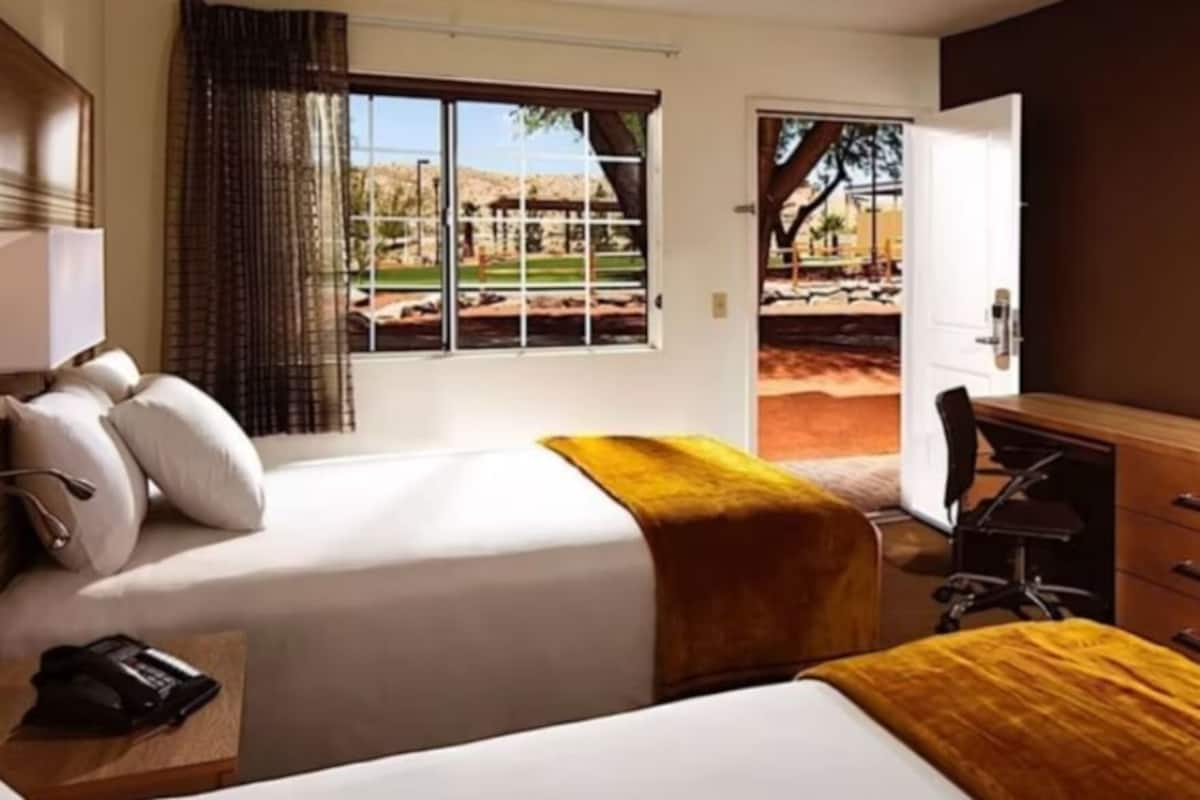 Habitación de hotel con dos camas, un escritorio, una silla, un televisor y una ventana con vistas a un parque.