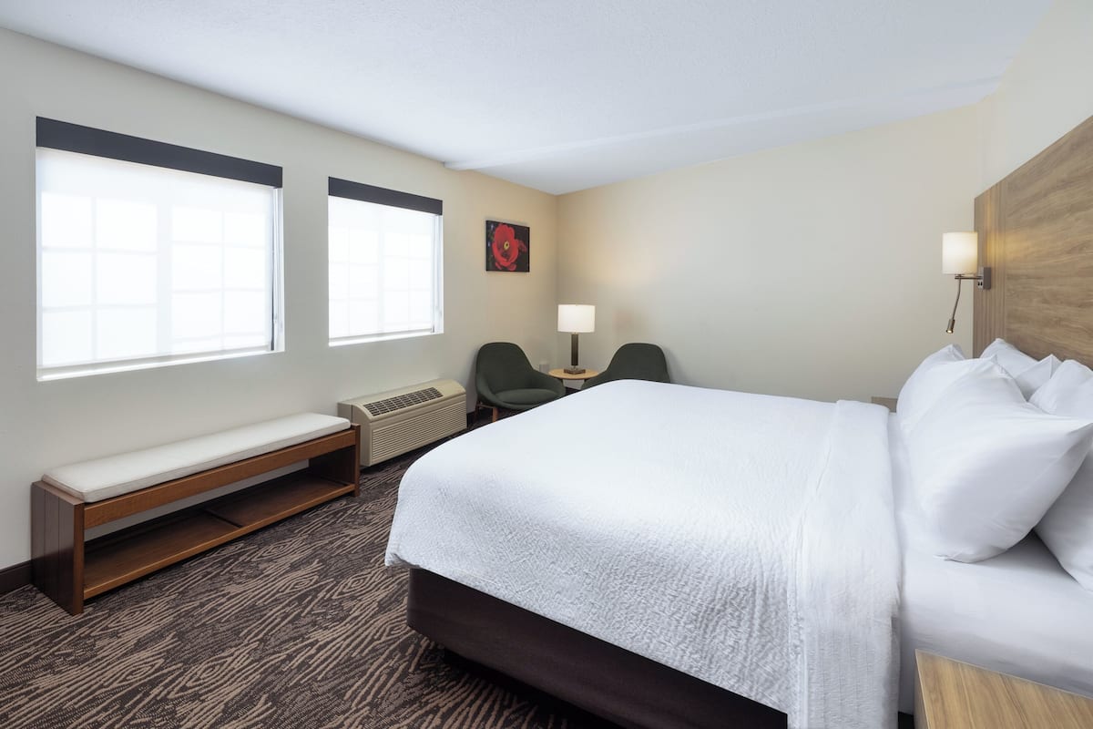 A hotel room with a large bed, a bench, two chairs, a small table, and a wall-mounted lamp.
