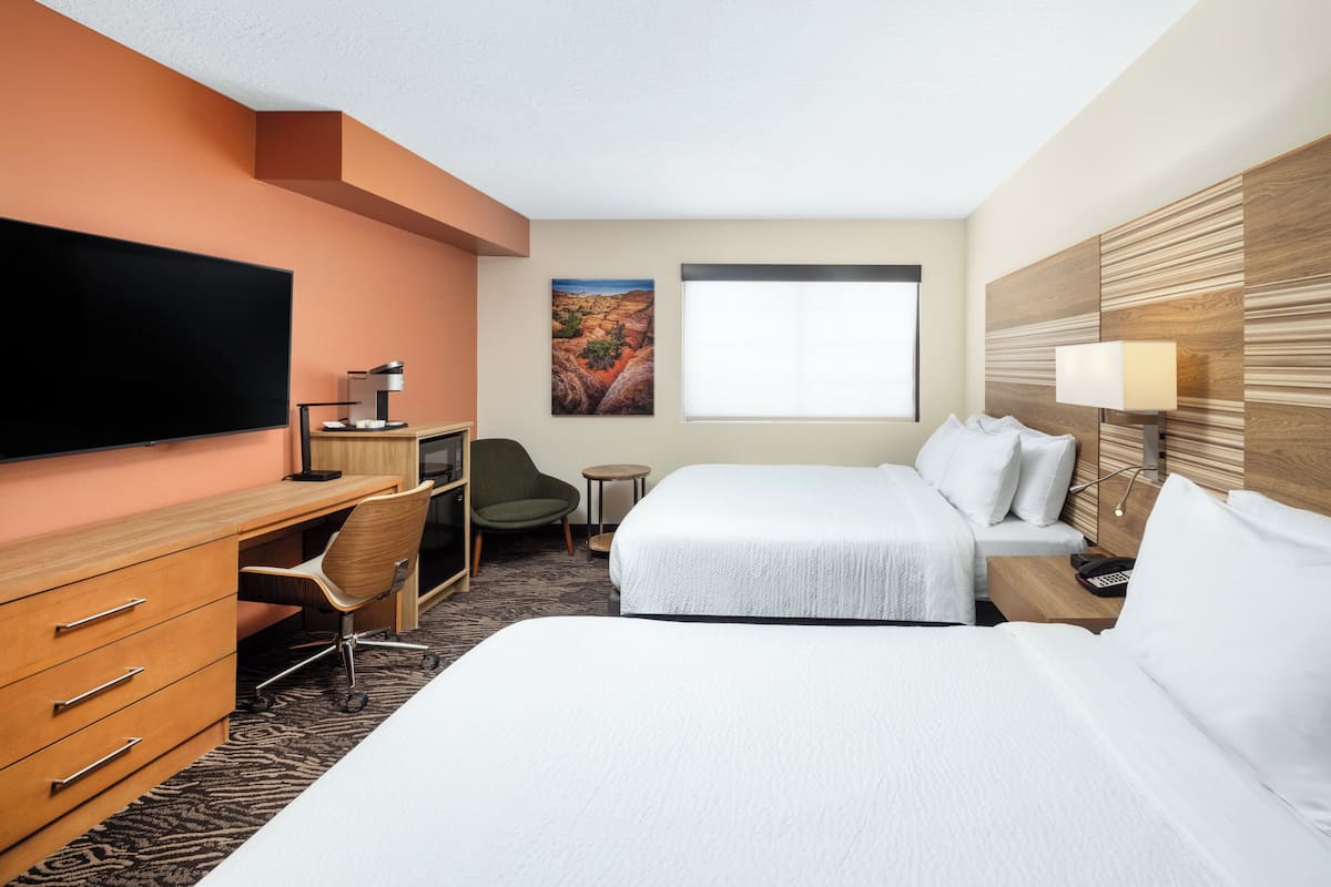 A hotel room with a large flat-screen TV, a desk with drawers, a chair, two beds with white bedding, a small round table, and a window with a view of a landscape.