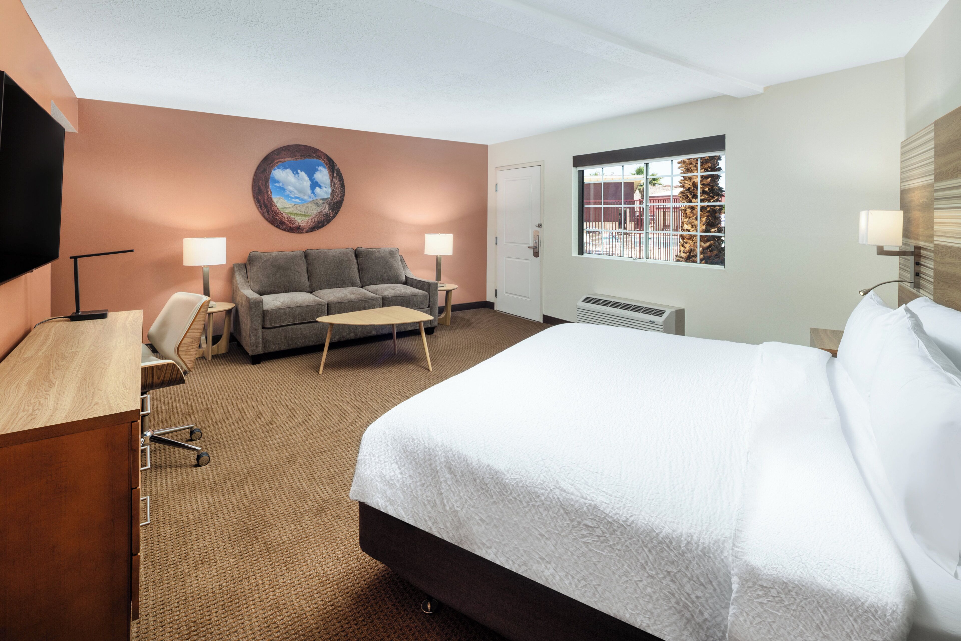 A hotel room with a bed, a sofa, a desk, a chair, two lamps, a painting, a window, and a door.