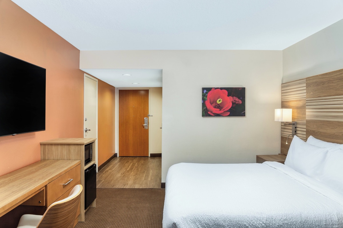 A hotel room with a bed, a desk with a chair, a television, a microwave, and a door leading to another room.