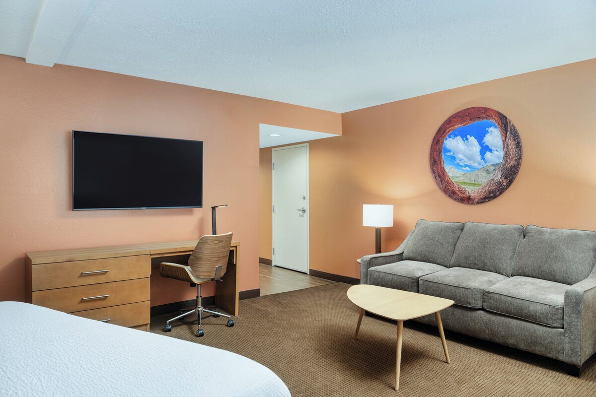 A hotel room with a bed, a desk, a grey sofa, a small table, a TV, and a door leading to another room.