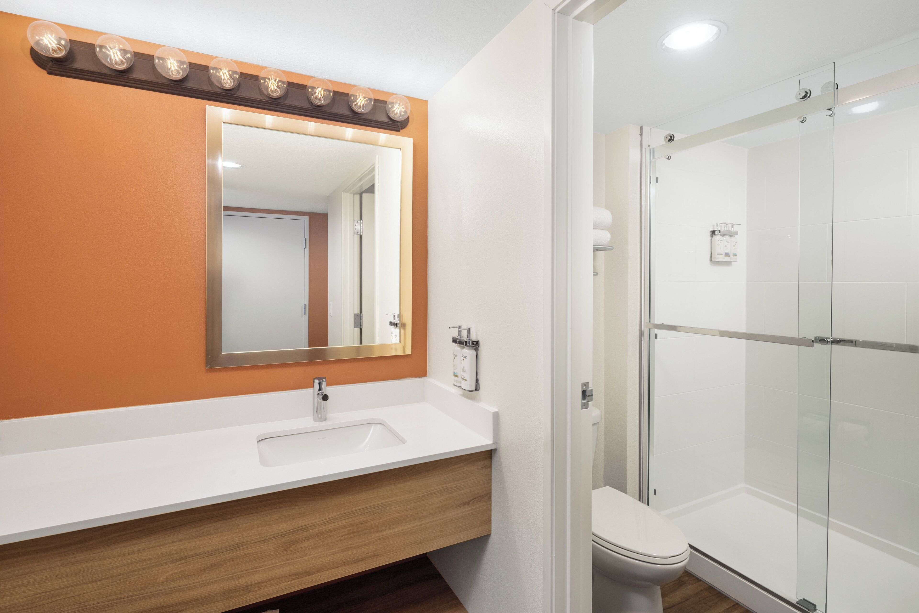 A bathroom with a white sink, a mirror with lights, an orange wall, and a glass shower enclosure.