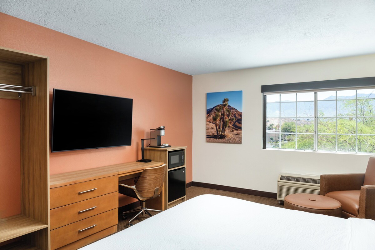 A hotel room with a bed, a desk with a flat-screen TV, a chair, a small kitchenette, and a window with a view of the outdoors.