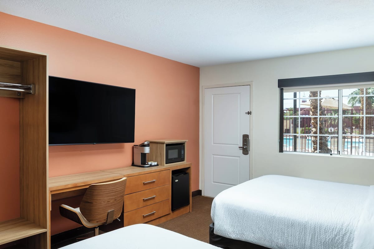 A hotel room with a wooden desk, a television, a microwave, a small refrigerator, a bed, and a window with a view of a pool and palm trees.
