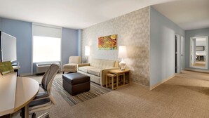A hotel room with a sofa, armchair, ottoman, table, and lamps. - Home2 Suites by Hilton Hasbrouck Heights (Hasbrouck Heights)