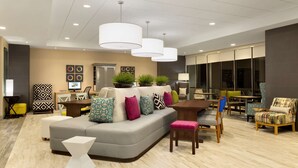 A modern living room with a grey sectional sofa, multiple chairs, a coffee table, and a dining area with a table and chairs. There are potted plants and artwork on the walls. - Home2 Suites by Hilton Hasbrouck Heights (Hasbrouck Heights)