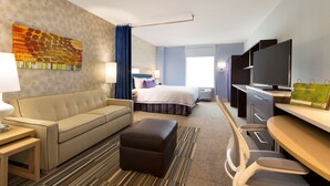 A hotel room with a bed, a sofa, a desk, a television, and a painting on the wall. - Home2 Suites by Hilton Hasbrouck Heights (Hasbrouck Heights)