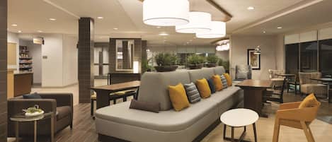 En modern hotellobby med en grå soffa, ett runt bord och en trästol. I bakgrunden finns krukväxter och ett stort fönster.