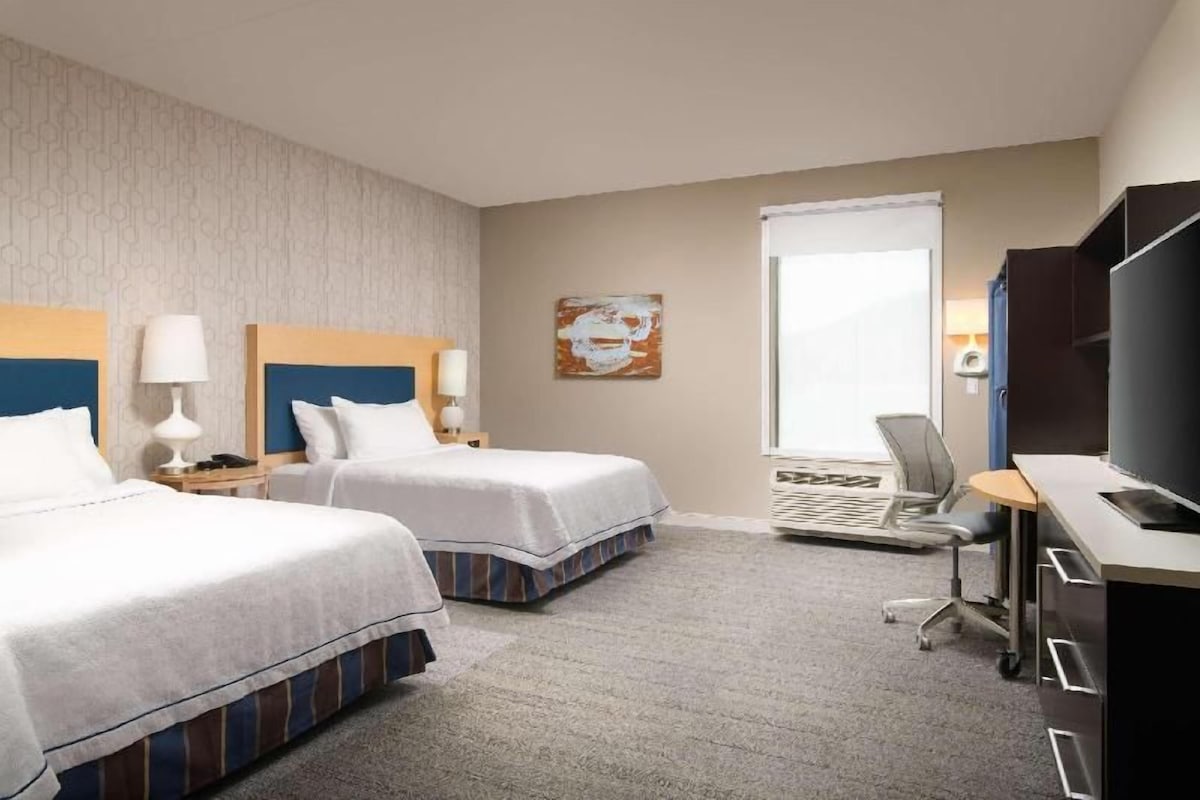 Quarto de hotel com duas camas, uma escrivaninha com cadeira, televisão e janela.