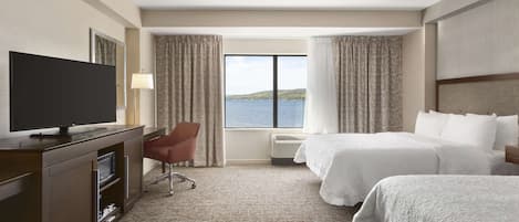 Une chambre d’hôtel dotée d’une grande télévision à écran plat, d’un bureau avec une chaise, de deux lits, d’une vue sur l’eau et d’une fenêtre avec des rideaux.
