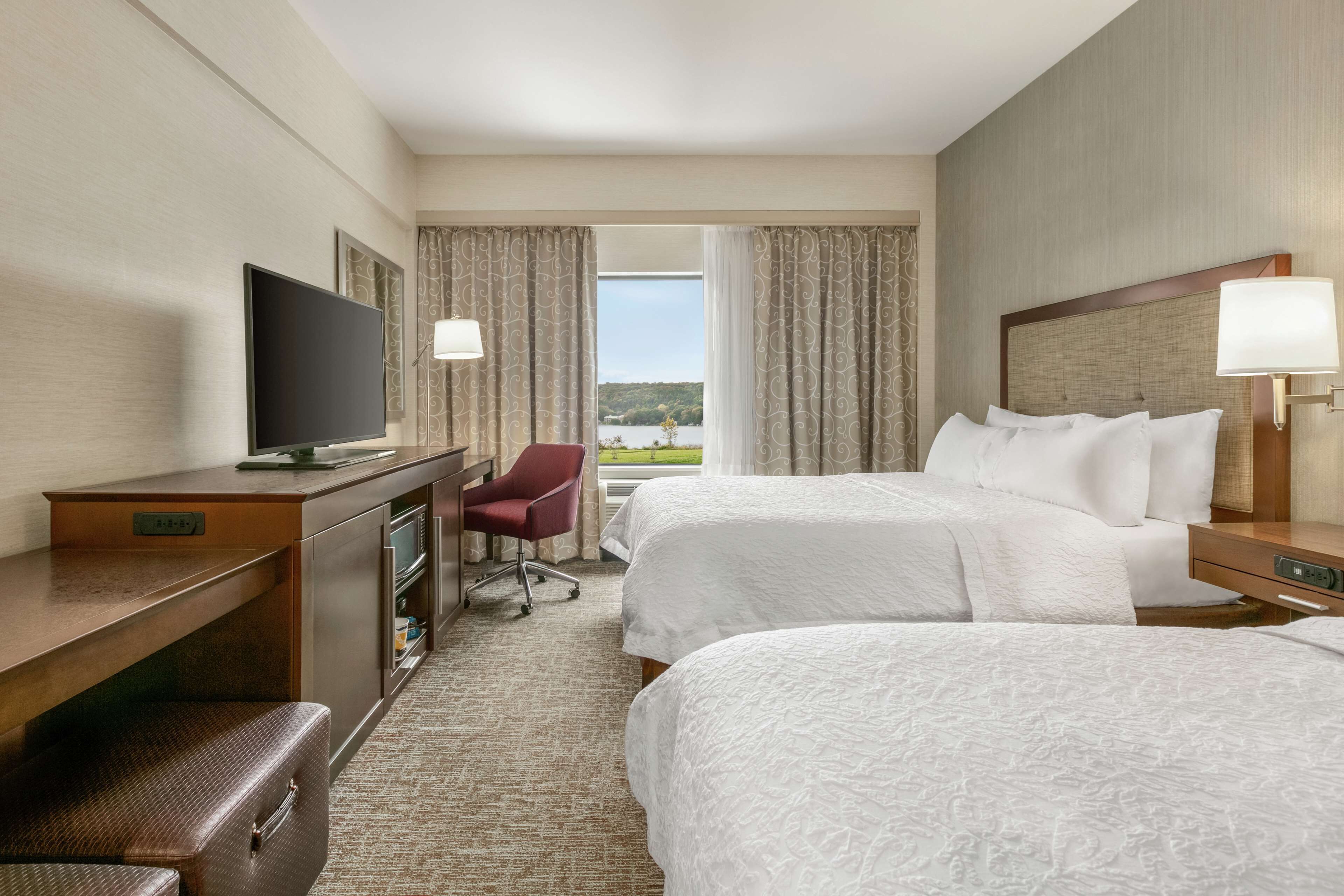 A hotel room with two beds, a desk with a TV, a chair, and a view of the outdoors.