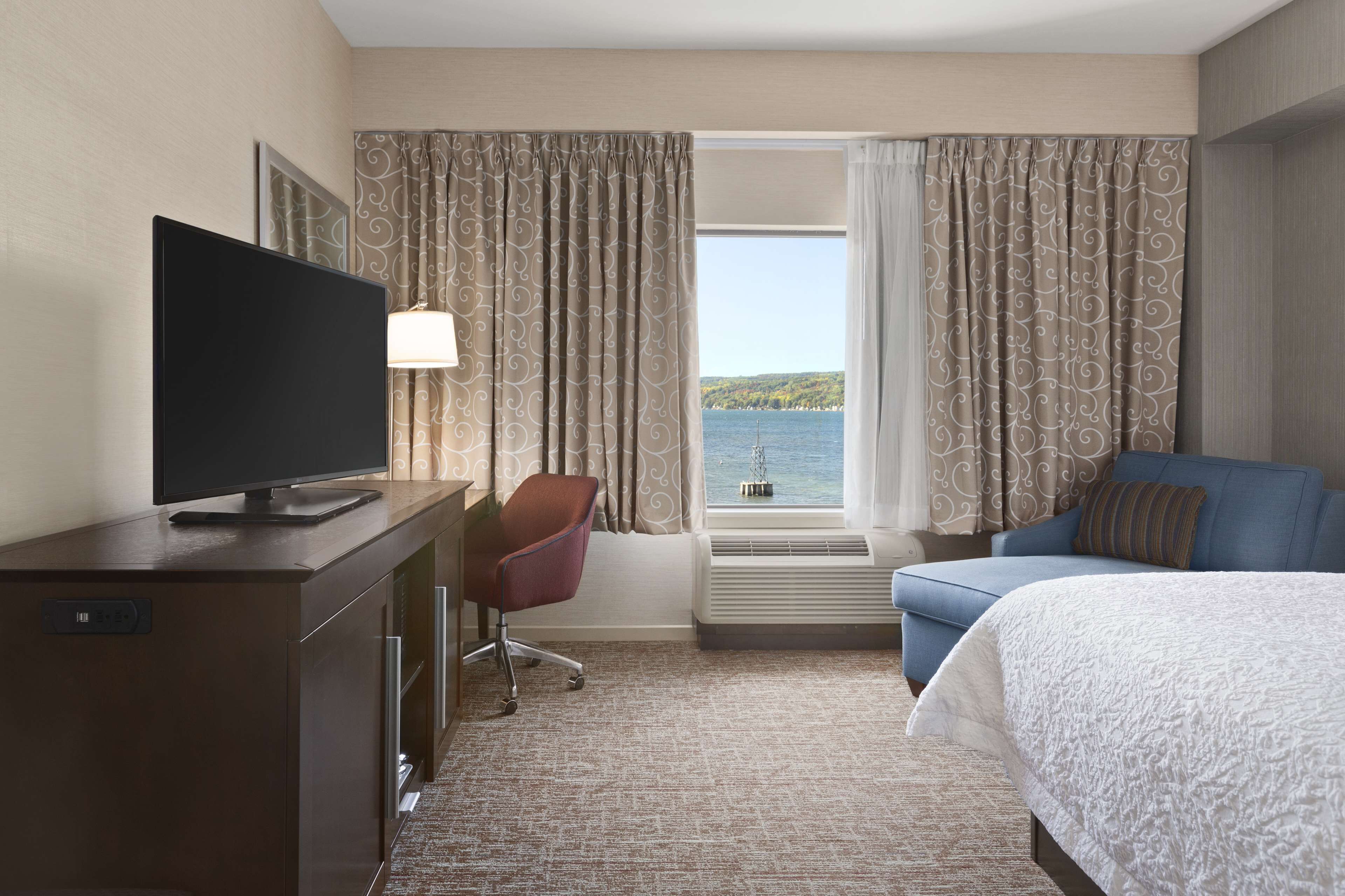 A hotel room with a bed, a desk with a TV, a chair, a window with curtains, and a view of the sea.