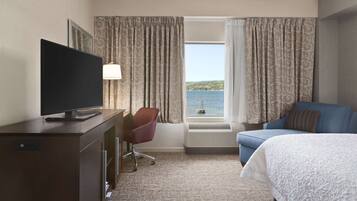 Habitación de hotel con cama, escritorio con televisor, silla, ventana con cortinas y vista al mar.