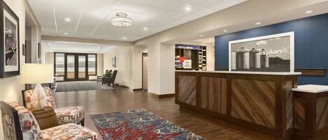 A hotel lobby with a reception desk, patterned chairs, a large rug, and a view of the outdoors.