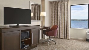 A hotel room with a TV on a wooden cabinet, a desk with a lamp, a chair, and a large window with curtains.