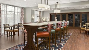 A spacious dining area with a long table and multiple chairs, overhead lighting, and large windows with curtains.