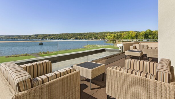 A balcony with wicker furniture, a glass table, and a view of a lake and forest.