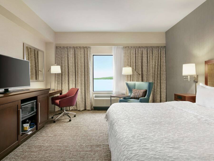 A hotel room with a large bed, a desk with a flat-screen TV, a red office chair, a blue armchair, and a view of the outdoors through a window.