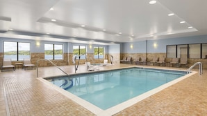 A large indoor swimming pool with clear blue water, surrounded by tiled flooring and walls. There are multiple seating areas with chairs and tables, and large windows offering views of the outdoors.