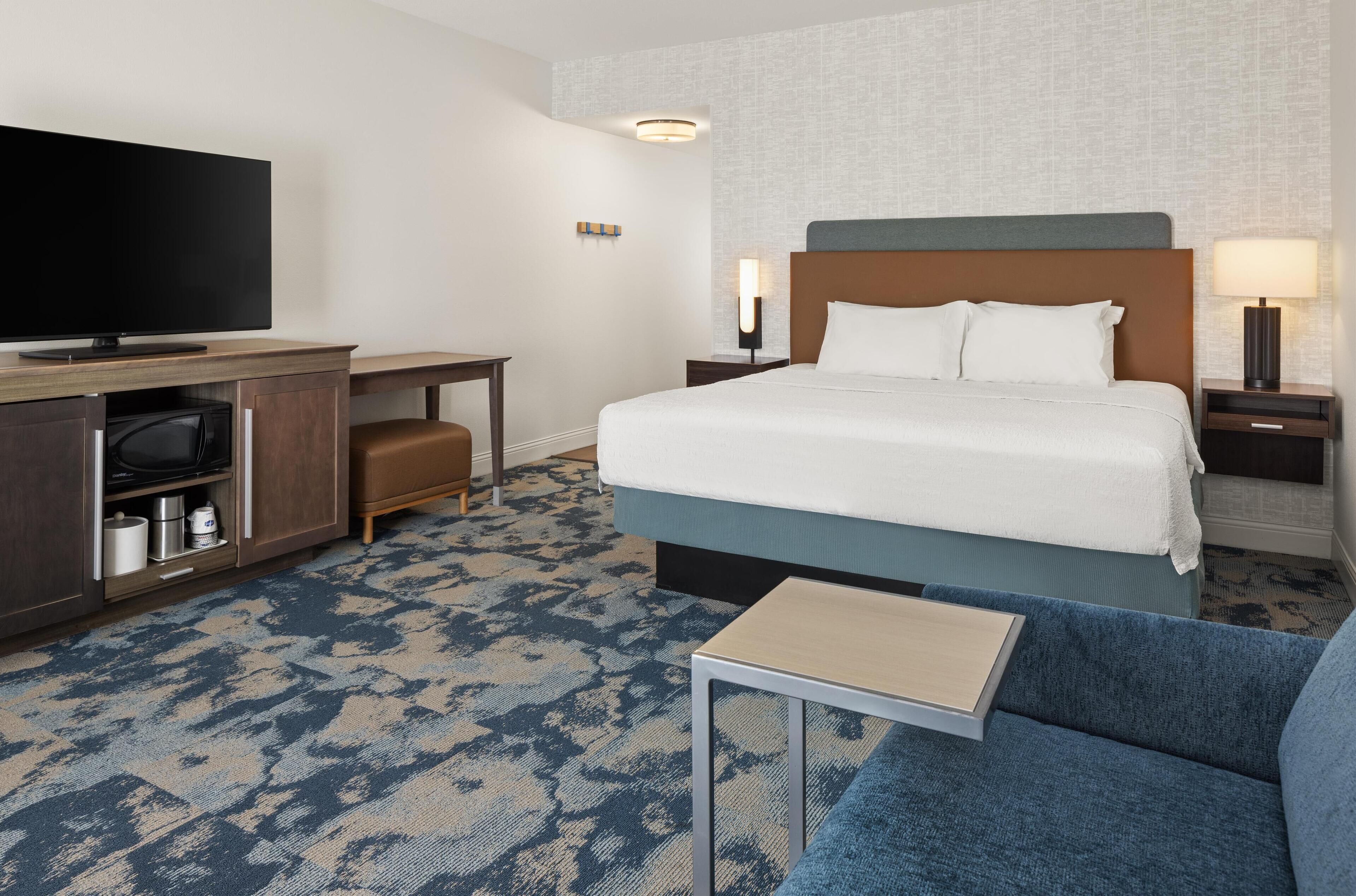 A hotel room with a bed, a TV, a desk, a lamp, and a sofa.