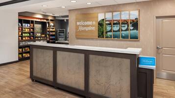 Una recepción de hotel con un cartel que dice “Welcome to Hampton”, estantes con aperitivos y un cartel de “Hilton Honors”.