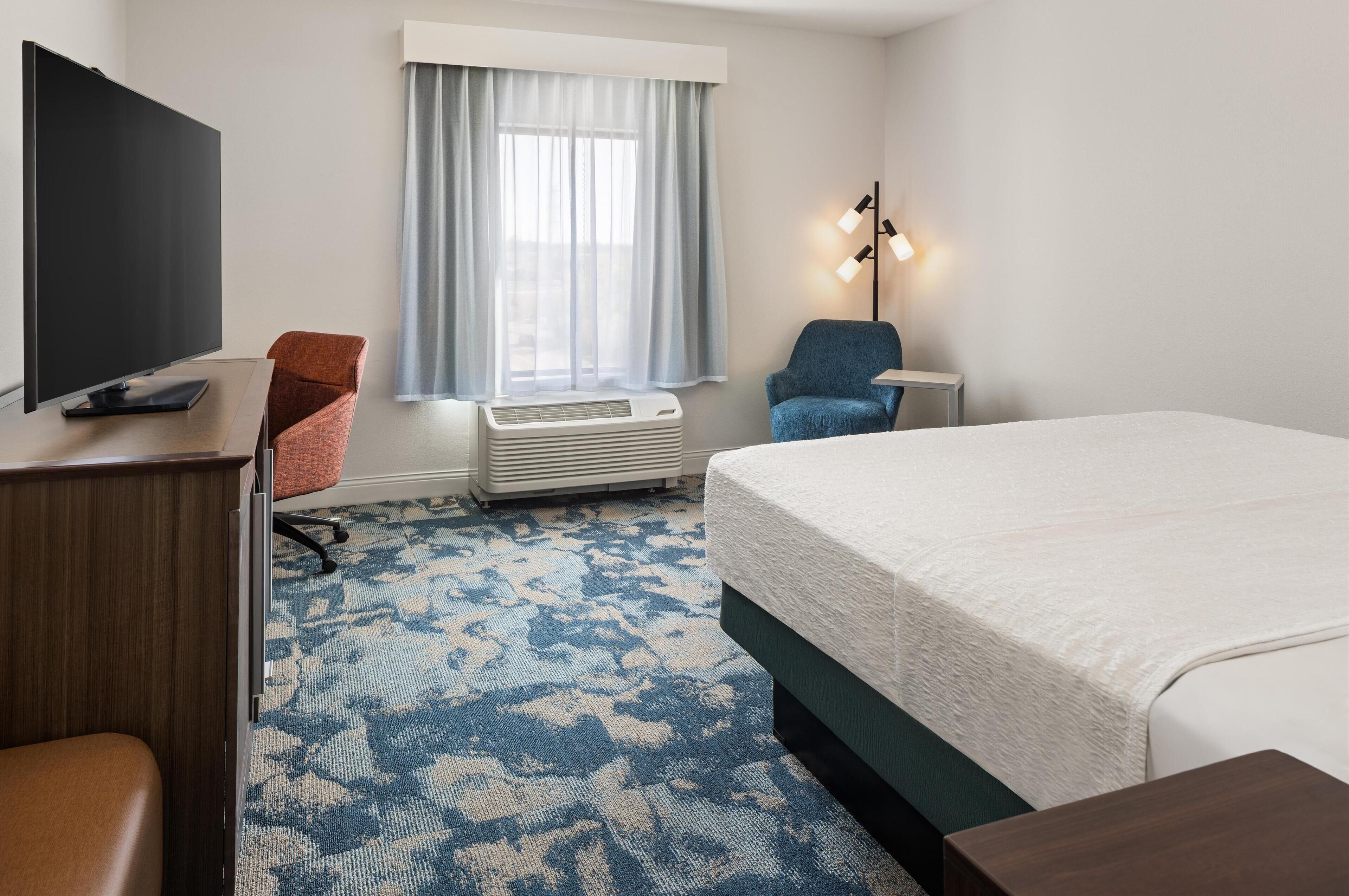 Habitación de hotel con cama, escritorio, televisor de pantalla plana, silla, lámpara de pie y ventana con cortinas.
