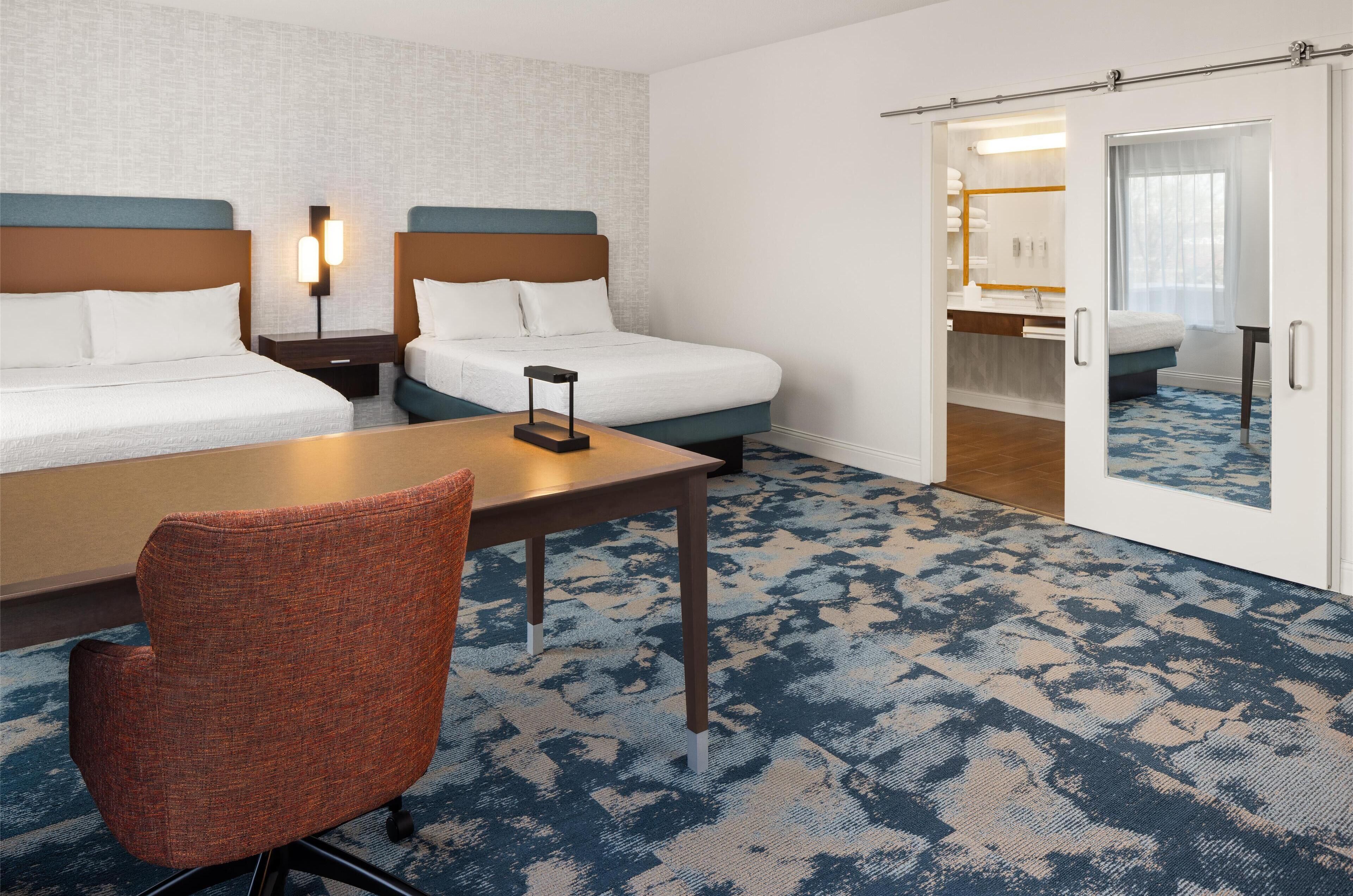 Habitación de hotel con dos camas, un escritorio, una silla y un baño visible a través de una puerta abierta.