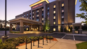 Um hotel Hampton Inn com fachada moderna, entrada coberta e estacionamento com suportes para bicicletas.