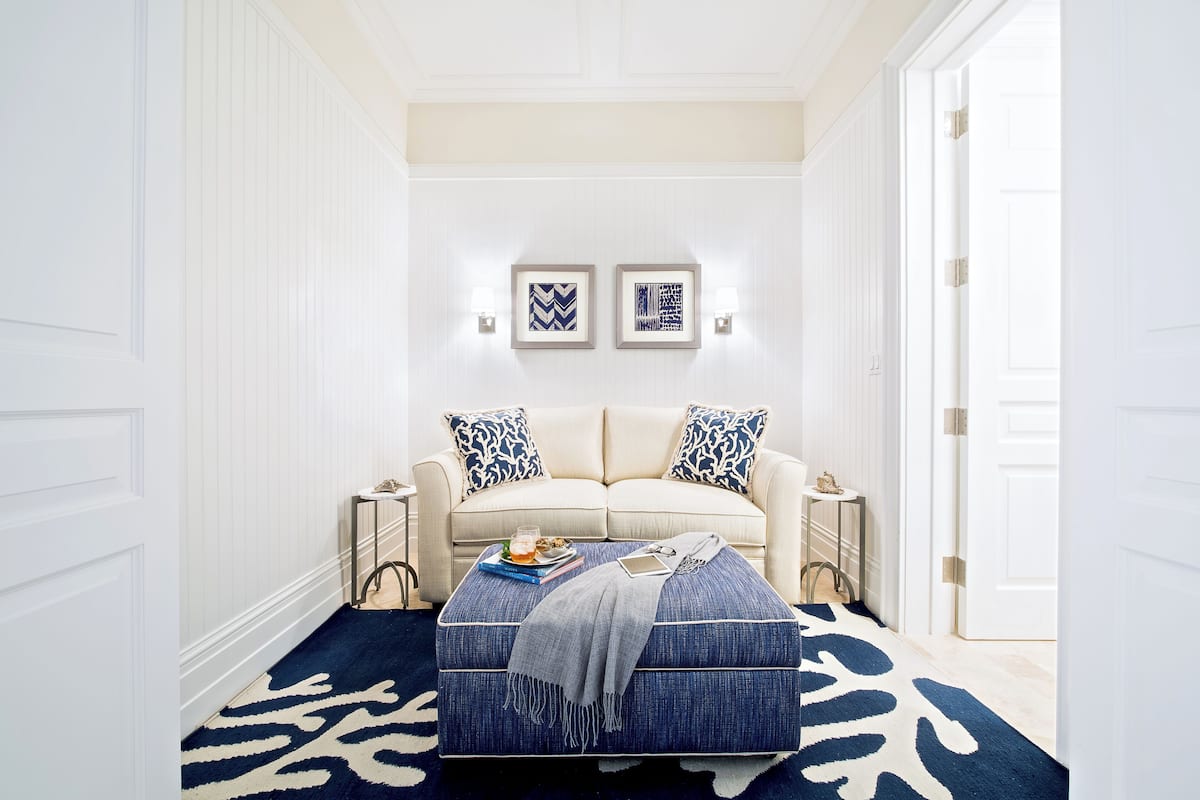 Un pasillo con una puerta blanca, una pared blanca, un tapete con diseño azul y blanco, un sofá blanco con almohadones azul y blanco, y dos cuadros en la pared.