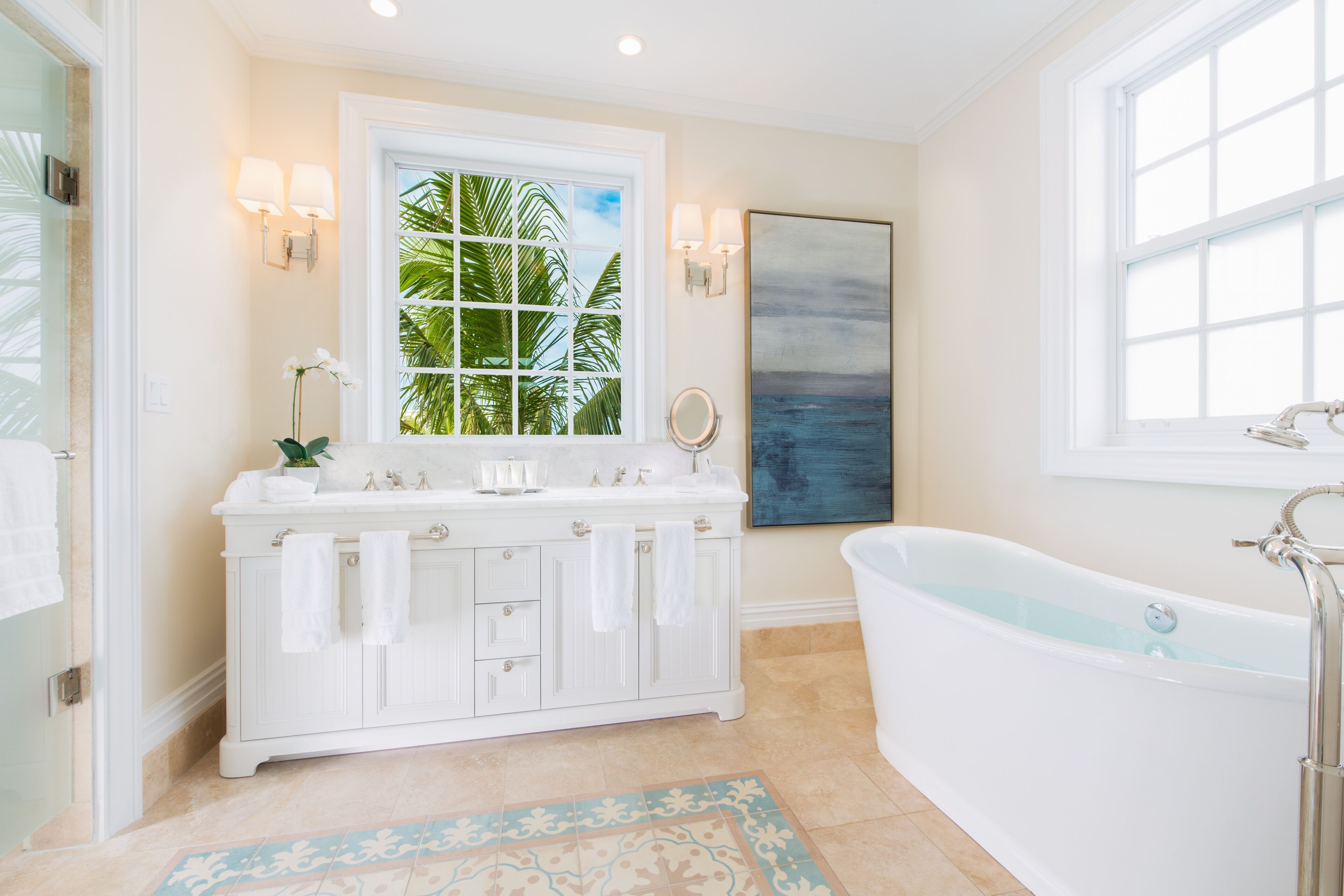 A bathroom with a white vanity, a bathtub, a window with a view of palm trees, and a painting on the wall.